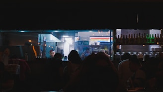 A dimly lit kitchen scene with a glass window revealing several people, including a chef wearing a cap. The kitchen is busy with various equipment and utensils visible. Outside the window, silhouettes of people can be seen in what appears to be a restaurant or bar setting. Shelves of bottles and jars are noticeable in the foreground.
