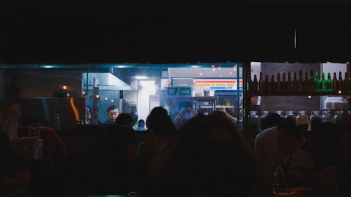 A dimly lit kitchen scene with a glass window revealing several people, including a chef wearing a cap. The kitchen is busy with various equipment and utensils visible. Outside the window, silhouettes of people can be seen in what appears to be a restaurant or bar setting. Shelves of bottles and jars are noticeable in the foreground.