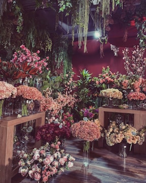 The image depicts a lush floral shop interior filled with a variety of vibrant flowers arranged in glass vases. There are wooden tables displaying colorful blooms such as hydrangeas, peonies, and roses. Hanging greenery decorates the ceiling, adding to the dense, botanical atmosphere. Price tags attached to some floral arrangements suggest a retail setting.