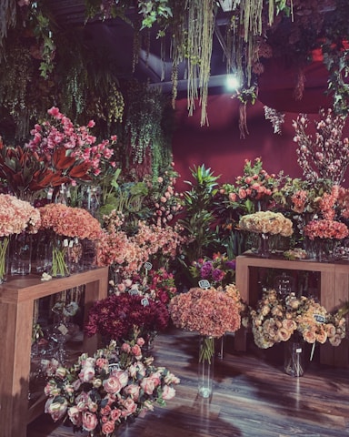 The image depicts a lush floral shop interior filled with a variety of vibrant flowers arranged in glass vases. There are wooden tables displaying colorful blooms such as hydrangeas, peonies, and roses. Hanging greenery decorates the ceiling, adding to the dense, botanical atmosphere. Price tags attached to some floral arrangements suggest a retail setting.