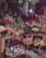 The image depicts a lush floral shop interior filled with a variety of vibrant flowers arranged in glass vases. There are wooden tables displaying colorful blooms such as hydrangeas, peonies, and roses. Hanging greenery decorates the ceiling, adding to the dense, botanical atmosphere. Price tags attached to some floral arrangements suggest a retail setting.