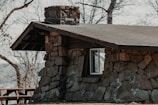 A small cabin with a stone chimney emitting smoke, set against a mountain backdrop.