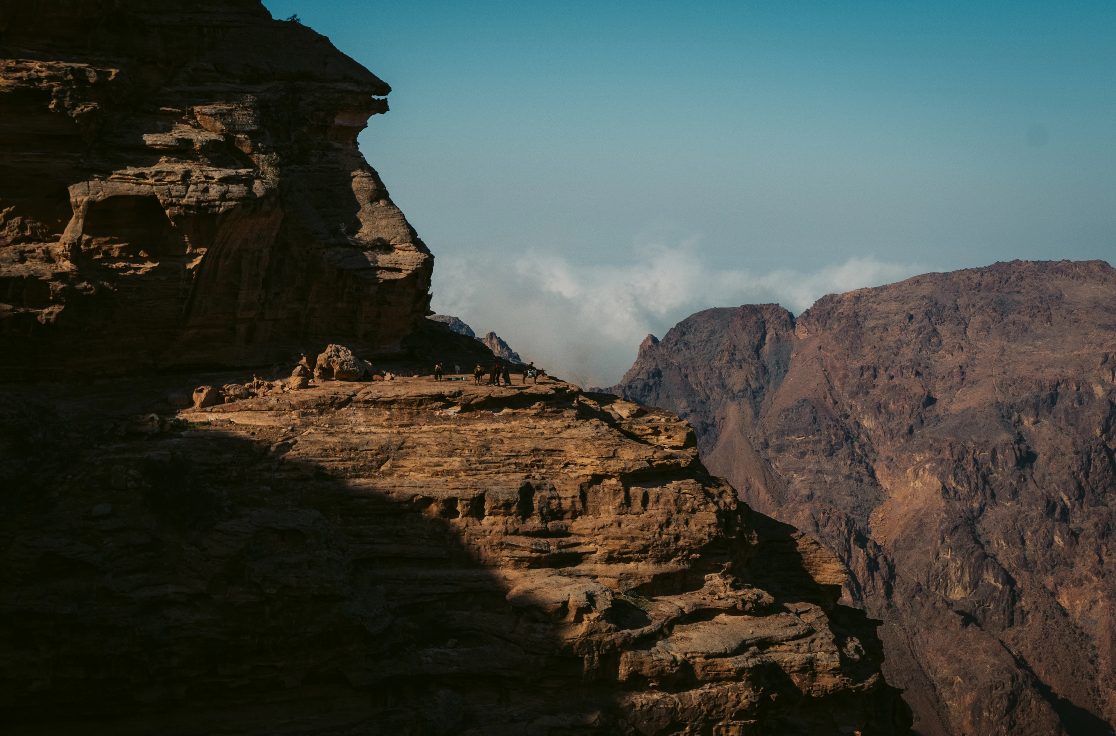 A high sandstone promontory at Petra with a handful of tiny figures standing along its sunlit rim, sheer layered cliffs dropping into shadow and the pale mountains of the Jordanian rift stacked behind under a deep blue sky.