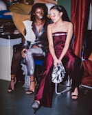 Two women are sitting in a backstage or studio environment, possibly preparing for a fashion show or event. The one on the left is wearing a metallic silver and black outfit with a unique textured shoulder design. The woman on the right is in a deep burgundy outfit and wearing red tinted sunglasses. Their surroundings have a utilitarian feel, with materials and sewing equipment visible in the background.