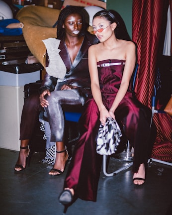 Two women are sitting in a backstage or studio environment, possibly preparing for a fashion show or event. The one on the left is wearing a metallic silver and black outfit with a unique textured shoulder design. The woman on the right is in a deep burgundy outfit and wearing red tinted sunglasses. Their surroundings have a utilitarian feel, with materials and sewing equipment visible in the background.