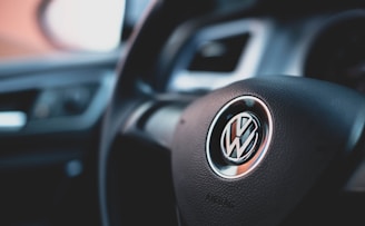 A close-up of a car steering wheel featuring a Volkswagen logo in the center. The interior of the car is visible with parts of the dashboard and vent in the background.