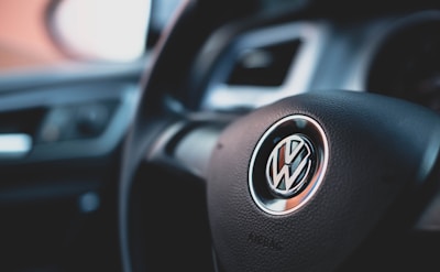 A close-up of a car steering wheel featuring a Volkswagen logo in the center. The interior of the car is visible with parts of the dashboard and vent in the background.
