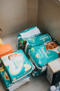 Several packages of Pampers baby diapers are arranged in an indoor setting. The packages are primarily turquoise and display images of smiling babies on the front. Various sizes of diaper packs are visible, indicating different quantities and designs.