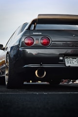 A close-up view of the rear side of a black Nissan car with a prominent spoiler. The car has circular red taillights and a visible exhaust pipe. The license plate is from Colorado, and small parts of other cars or people appear in the background.
