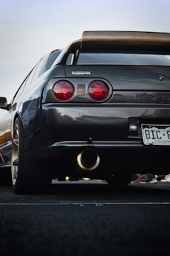 A close-up view of the rear side of a black Nissan car with a prominent spoiler. The car has circular red taillights and a visible exhaust pipe. The license plate is from Colorado, and small parts of other cars or people appear in the background.