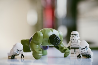 A group of LEGO figures includes a green, muscular figure in a dynamic pose surrounded by several small, white-armored figures. The scene is set against a blurred background that likely suggests an indoor environment.