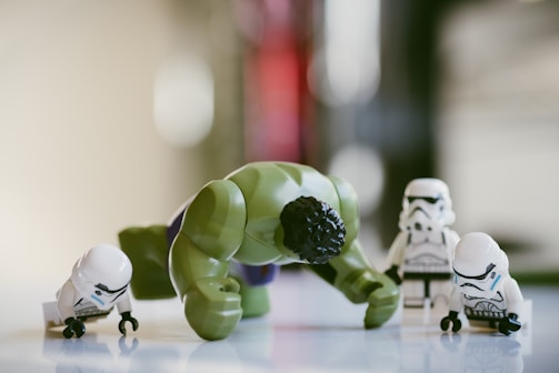 A group of LEGO figures includes a green, muscular figure in a dynamic pose surrounded by several small, white-armored figures. The scene is set against a blurred background that likely suggests an indoor environment.