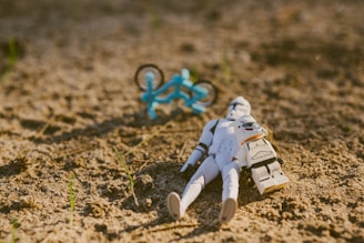 A toy stormtrooper figure lies on sandy ground with a small blue bicycle in the background. The lighting is warm, suggesting a sunny day, with a few green sprouts emerging from the sand.
