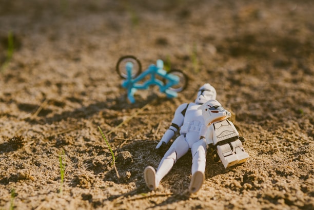 A toy stormtrooper figure lies on sandy ground with a small blue bicycle in the background. The lighting is warm, suggesting a sunny day, with a few green sprouts emerging from the sand.