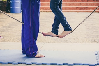Two individuals are practicing tightrope walking outdoors. One person is standing barefoot on a tightrope while another is positioned opposite, providing balance support. Both are wearing casual clothing with the person on the left in purple pants and the one balancing in jeans.