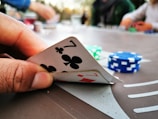 A close-up of a hand holding two playing cards, a seven of clubs and a seven of diamonds. In the background, there are poker chips in different colors, such as blue and green, scattered on a table. The image suggests a casual setting with people playing poker.