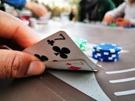 A close-up of a hand holding two playing cards, a seven of clubs and a seven of diamonds. In the background, there are poker chips in different colors, such as blue and green, scattered on a table. The image suggests a casual setting with people playing poker.