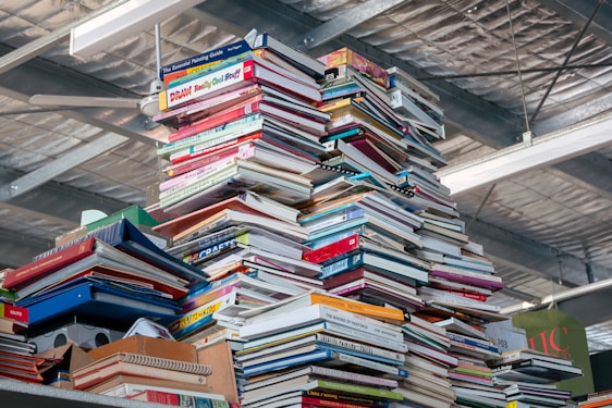 A large stack of colorful books, magazines, and notebooks piled high, reaching toward a ceiling in an indoor setting. The background features industrial metal beams and lights.