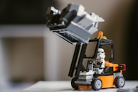 A LEGO construction setup featuring a mini stormtrooper figure operating a small, orange and black forklift model. The forklift is lifting a gray structure, and a small amber light is mounted on top of the vehicle. The background is blurred, creating a focus on the LEGO scene.