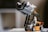 A LEGO construction setup featuring a mini stormtrooper figure operating a small, orange and black forklift model. The forklift is lifting a gray structure, and a small amber light is mounted on top of the vehicle. The background is blurred, creating a focus on the LEGO scene.