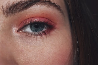 A close-up of a person's eye with blue iris, framed by bold eyebrows. The eyelid is adorned with a red-orange eyeshadow, highlighting the eye's features.