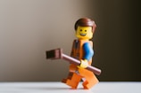 A Lego figurine with a smiling face, wearing an orange construction outfit and holding a brown stick. The figurine has brown hair and is positioned against a blurred, neutral background.
