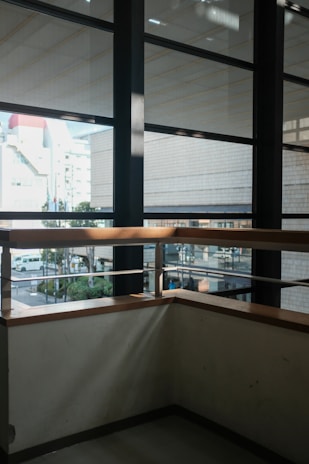 A modern interior view featuring large glass windows with black metal frames providing a view of an urban environment outside. The windows offer glimpses of buildings and a street with a few visible vehicles and trees. Inside, a corner space with light-colored walls and a wooden railing, where daylight creates subtle shadows and reflections.