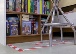 A bookshelf filled with a variety of science fiction and fantasy novels, including titles like 'The Iron Tower' and 'The War of Powers'. The books are organized neatly on wooden shelves. In the foreground, there is a metallic chair with visible legs, placed over a floor with white and red hazard tape markings.