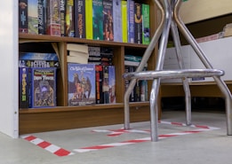 A bookshelf filled with a variety of science fiction and fantasy novels, including titles like 'The Iron Tower' and 'The War of Powers'. The books are organized neatly on wooden shelves. In the foreground, there is a metallic chair with visible legs, placed over a floor with white and red hazard tape markings.