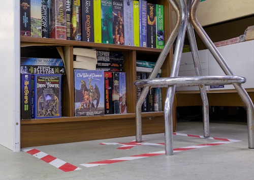 A bookshelf filled with a variety of science fiction and fantasy novels, including titles like 'The Iron Tower' and 'The War of Powers'. The books are organized neatly on wooden shelves. In the foreground, there is a metallic chair with visible legs, placed over a floor with white and red hazard tape markings.