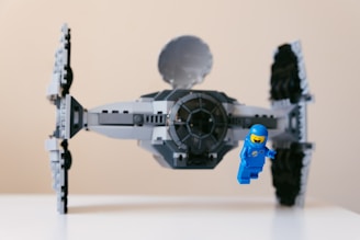 A detailed LEGO model of a spacecraft is positioned prominently, featuring intricate design elements and a grey color scheme. A small blue LEGO astronaut figure appears alongside, seemingly floating in space, adding a playful and imaginative element to the scene.