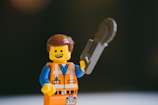A LEGO figure with a smiling expression holds a grey tool resembling a saw. The figure is dressed in an orange and blue outfit with a utility vest. The background is dark, making the figure stand out.