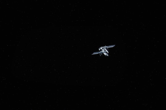 A space station is positioned slightly off-center against a backdrop of a star-filled sky. The station is illuminated, showcasing its solar panels and structural complexity, while the majority of the frame is dominated by the vast darkness of space, dotted with small distant stars.