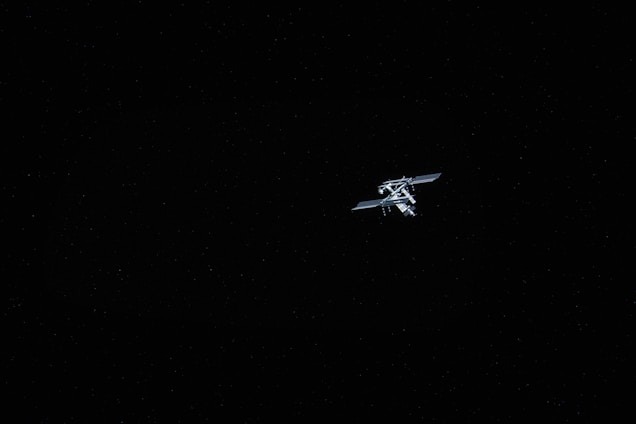 A space station is positioned slightly off-center against a backdrop of a star-filled sky. The station is illuminated, showcasing its solar panels and structural complexity, while the majority of the frame is dominated by the vast darkness of space, dotted with small distant stars.