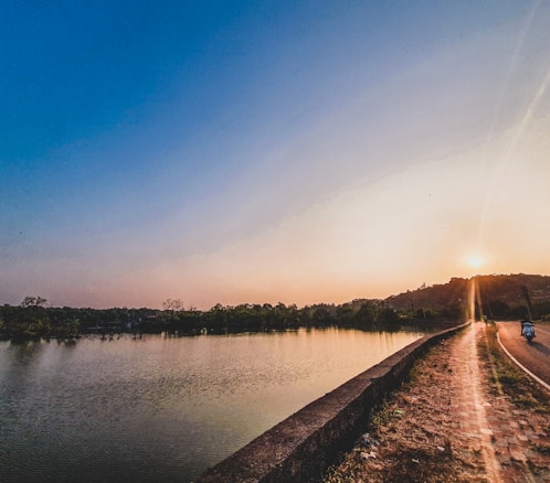 A peaceful landscape featuring a serene body of water on the left and a paving walkway beside it leading towards a distant, softly glowing sunset. The sky transitions from a deep blue at the top to warm hues near the horizon. Hills and silhouettes of trees border the water, adding a natural, tranquil atmosphere.