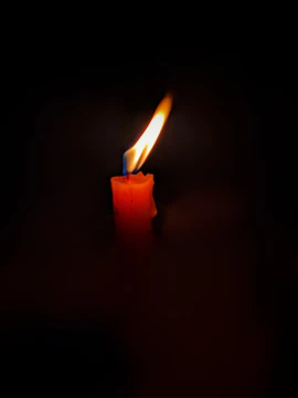 A brightly burning candle in the dark, casting a warm glow with a vivid flame on top of a red wax pillar.