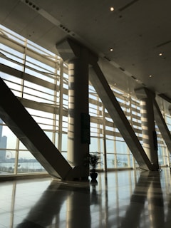 A spacious modern interior with large floor-to-ceiling windows casting geometric patterns of light and shadow on the glossy floor. The space includes tall structural columns and a view of an urban landscape outside, with a glimpse of a Ferris wheel in the background. Indoor plants add a touch of greenery.
