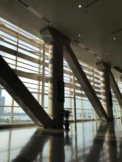A spacious modern interior with large floor-to-ceiling windows casting geometric patterns of light and shadow on the glossy floor. The space includes tall structural columns and a view of an urban landscape outside, with a glimpse of a Ferris wheel in the background. Indoor plants add a touch of greenery.