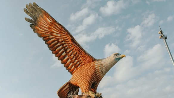 A large eagle statue with its wings spread wide against a backdrop of a partly cloudy sky. The statue appears highly detailed, showcasing the texture of the feathers and the intensity of the eagle's gaze. A tall metal pole with lights is visible on the right side.