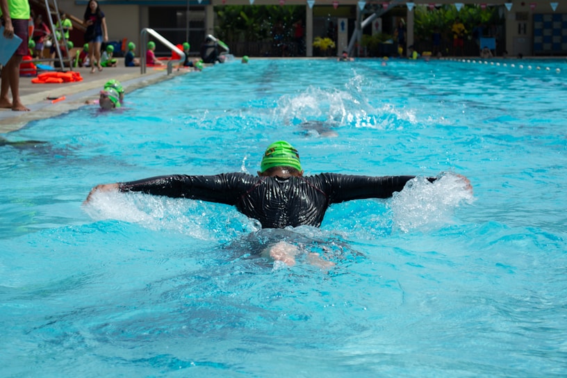 A person is swimming in a pool wearing a black wetsuit and a bright green swim cap. The swimmer is performing the butterfly stroke, with arms outstretched and water splashing around. In the background, several people in colorful swim caps are also visible, and there are onlookers and a poolside area with various items such as towels and swim gear.