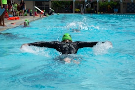 A person is swimming in a pool wearing a black wetsuit and a bright green swim cap. The swimmer is performing the butterfly stroke, with arms outstretched and water splashing around. In the background, several people in colorful swim caps are also visible, and there are onlookers and a poolside area with various items such as towels and swim gear.