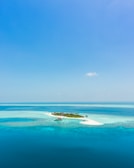 A small tropical island surrounded by crystal clear turquoise waters and white sandy beaches. The island is lush with green vegetation and appears isolated under a bright blue sky.