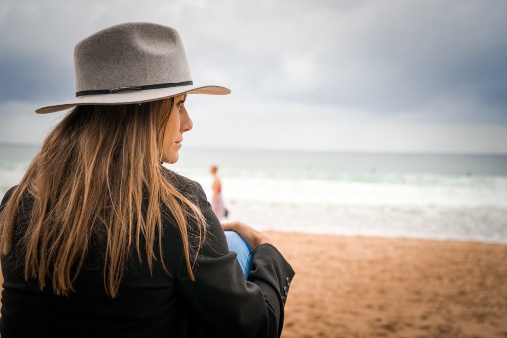 Donna in camicia nera a maniche lunghe e cappello Fedora marrone seduta sulla spiaggia durante il giorno
