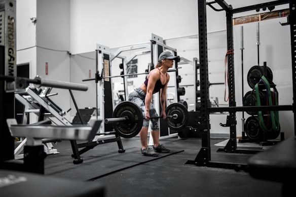 A person is in a gym setting performing a deadlift with a barbell. The individual is wearing athletic clothing, including a cap, a black top, and patterned leggings. The gym is equipped with various weightlifting and exercise machines and equipment, such as a squat rack, weight plates, and resistance bands.