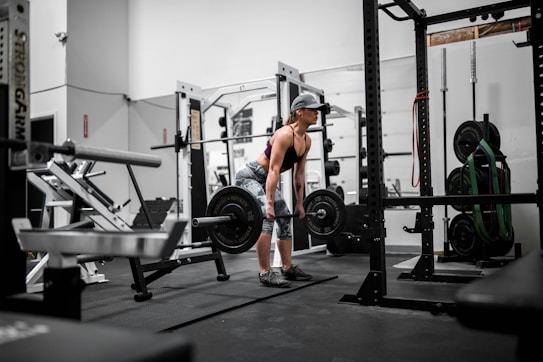 A person is in a gym setting performing a deadlift with a barbell. The individual is wearing athletic clothing, including a cap, a black top, and patterned leggings. The gym is equipped with various weightlifting and exercise machines and equipment, such as a squat rack, weight plates, and resistance bands.