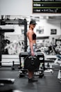 A woman is standing in a gym holding a heavy barbell plate marked with 'YORK 45'. She is wearing a cap, sports bra, and patterned leggings. The gym is equipped with various fitness machines and weights.