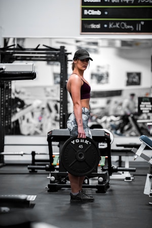A woman is standing in a gym holding a heavy barbell plate marked with 'YORK 45'. She is wearing a cap, sports bra, and patterned leggings. The gym is equipped with various fitness machines and weights.