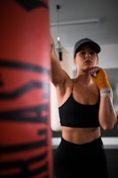 A person wearing a black sports bra and hat is in a gym setting, appearing focused and ready to punch a boxing bag. The individual has orange hand wraps and is standing in a confident pose. The background includes some gym equipment, indicating a training environment.