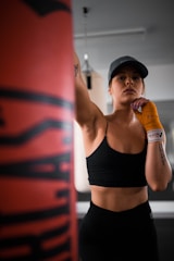 A person wearing a black sports bra and hat is in a gym setting, appearing focused and ready to punch a boxing bag. The individual has orange hand wraps and is standing in a confident pose. The background includes some gym equipment, indicating a training environment.