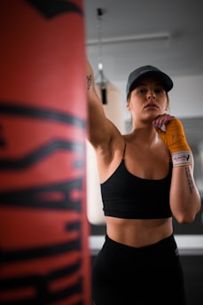 A person wearing a black sports bra and hat is in a gym setting, appearing focused and ready to punch a boxing bag. The individual has orange hand wraps and is standing in a confident pose. The background includes some gym equipment, indicating a training environment.